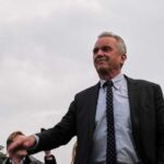 Robert F. Kennedy Jr. in a suit stands outdoors, cloudy sky background.