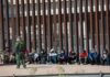 Border patrol officer watches group sitting by a fence.