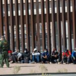 Border patrol officer watches group sitting by a fence.