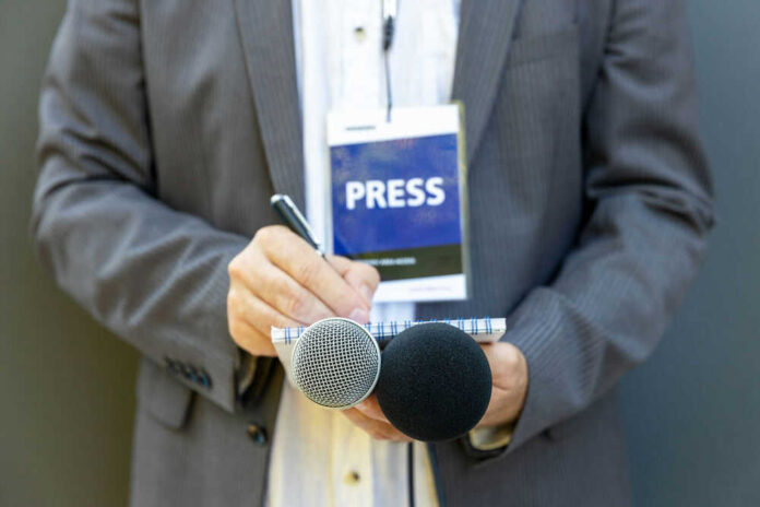Journalist with press badge, notebook, and microphones.
