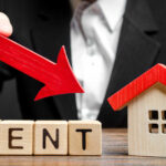 A business professional pointing a red arrow down towards the word 'RENT' made of wooden blocks next to a small house model