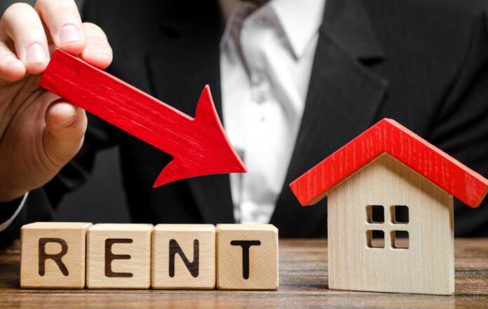 A business professional pointing a red arrow down towards the word 'RENT' made of wooden blocks next to a small house model