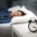 An alarm clock on a bedside table with a person sleeping in the background