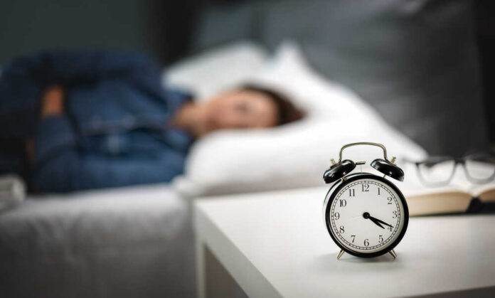 An alarm clock on a bedside table with a person sleeping in the background