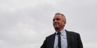Robert F. Kennedy Jr. in a suit stands outdoors, cloudy sky background.