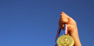 A hand holding a gold medal against a clear blue sky