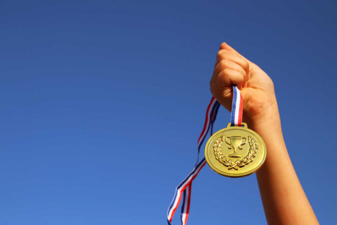 shutterstock_1111960664.jpg A hand holding a gold medal against a clear blue sky