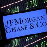 A hand holding a smartphone displaying the JPMorgan Chase logo in front of a stock market screen