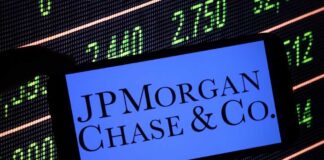 A hand holding a smartphone displaying the JPMorgan Chase logo in front of a stock market screen