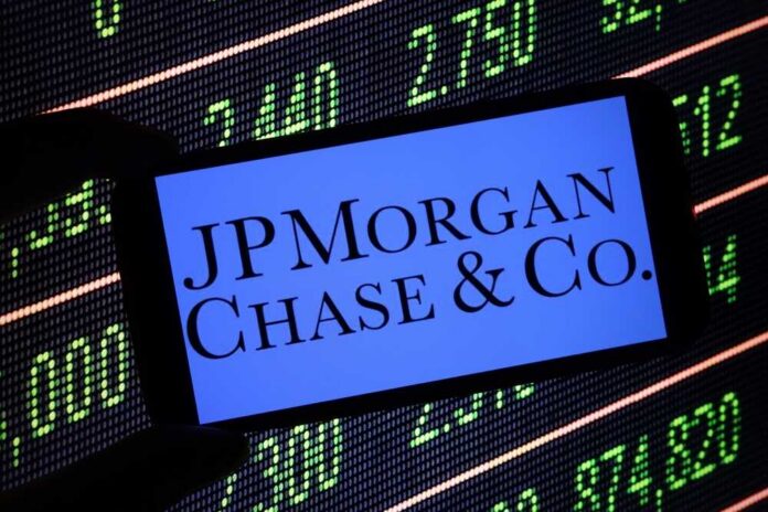 A hand holding a smartphone displaying the JPMorgan Chase logo in front of a stock market screen