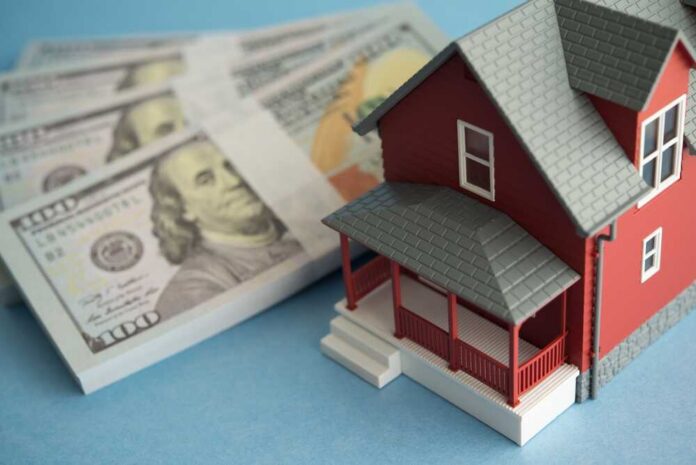 A model red house next to stacks of hundred-dollar bills