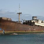 An abandoned, rusted ship stranded on the shore with a calm ocean in the background