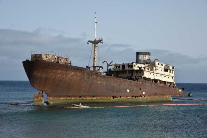 An abandoned, rusted ship stranded on the shore with a calm ocean in the background