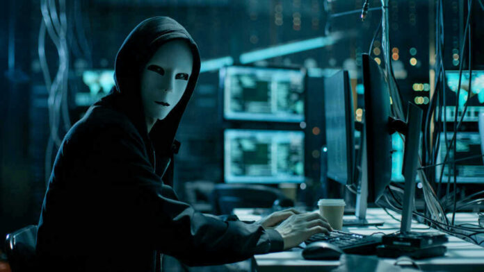 Person in hoodie and mask using computer, multiple screens.