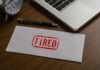 An envelope marked 'FIRED' on a wooden desk next to a clock and a laptop