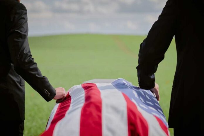 Two individuals standing beside a flag-draped casket in a green field