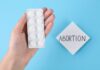 A hand holding a blister pack of white pills next to a note that says 'ABORTION'