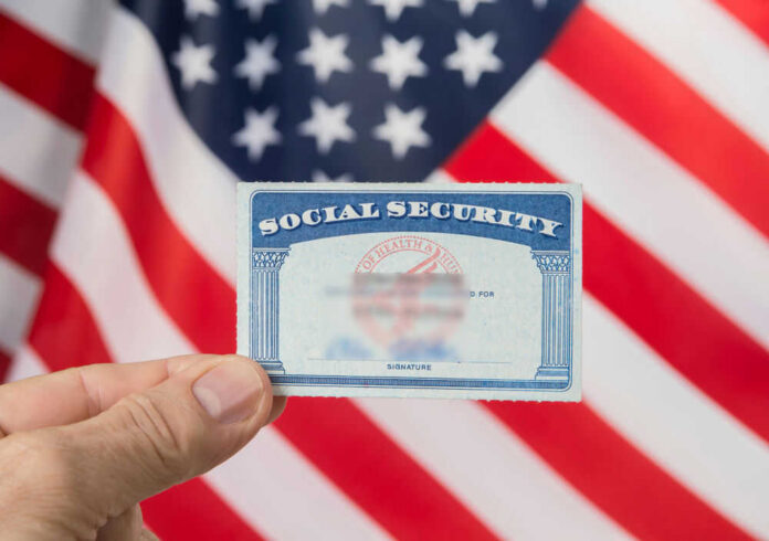 shutterstock_1125089156 Hand holding social security card, American flag background.