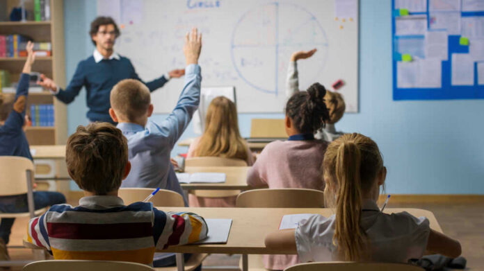 Students raising hands in a classroom with a teacher.