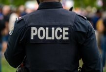 Body Found… But Is It Her?! Back view of a police officer wearing a uniform with 'POLICE' displayed