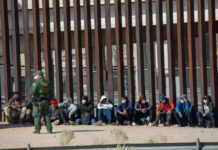 ICE Blitz Shocks — 10,000 Nabbed In Days Border patrol officer watches group sitting by a fence.