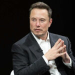 Zelenskyy’s Bold Nod To Elon Musk Man in suit, hands clasped, black background.
