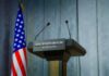 West Wing Whiplash: Press Boss Exits Press podium with the White House sign and an American flag in the background