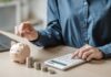 Person placing a coin into a piggy bank while using a calculator