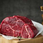 Mystery Beef Pulled — Crucial Details HERE! A raw cut of beef on a wooden board with herbs and a pepper grinder