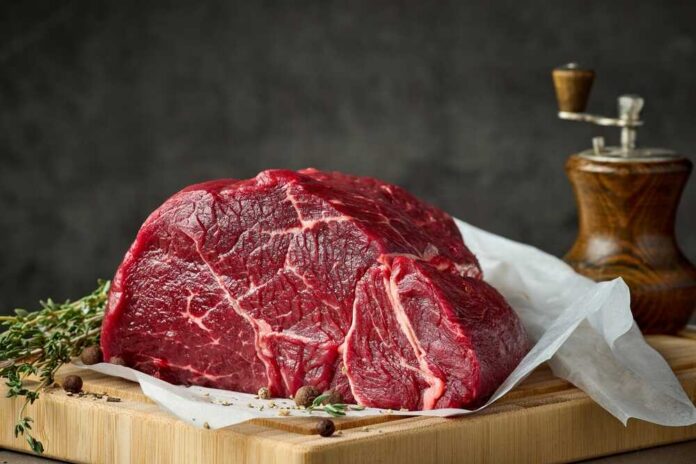 Fresh,Raw,Beef,Meat,With,Herbs,And,Spices,On,Wooden A raw cut of beef on a wooden board with herbs and a pepper grinder