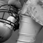 TRAGEDY: NFL’s Grim Math – One In Four Football player holding helmet, dirty uniform close-up.