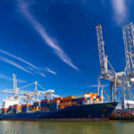 Pentagon’s DRONE Strike Sparks Iran Tensions Cargo ship docked at port with cranes unloading.
