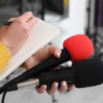 NOW: Car Bills Explode Journalist holding two microphones while writing in a notebook