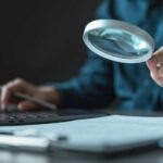 Rate Shock Slams THESE Americans Person using a magnifying glass while working on a laptop