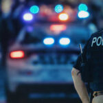 Deadly Block Party Chaos Police officer stands near patrol car with lights flashing.