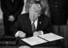 man in suit signs a document at a desk